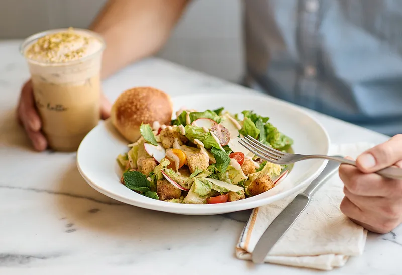 A person holds a fork, ready to eat a fresh salad with croutons and radishes. An iced coffee and a roll sit on the table next to them.