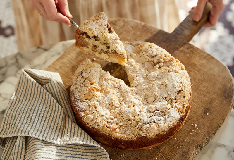 A person removes a slice from a nut-topped crumb cake on a wooden board, next to a striped cloth.