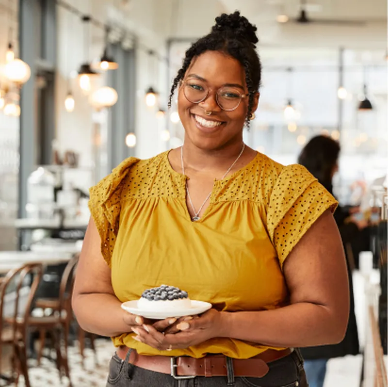 Smiling woman with glasses in a café holds a dessert topped with blueberries. She wears a mustard yellow top, standing in a well-lit space.
