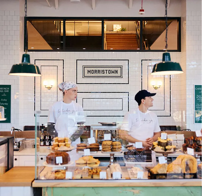 Two bakery staff behind a counter with pastries, under hanging lights. 'MORRISTOWN' sign on tiled wall in background.