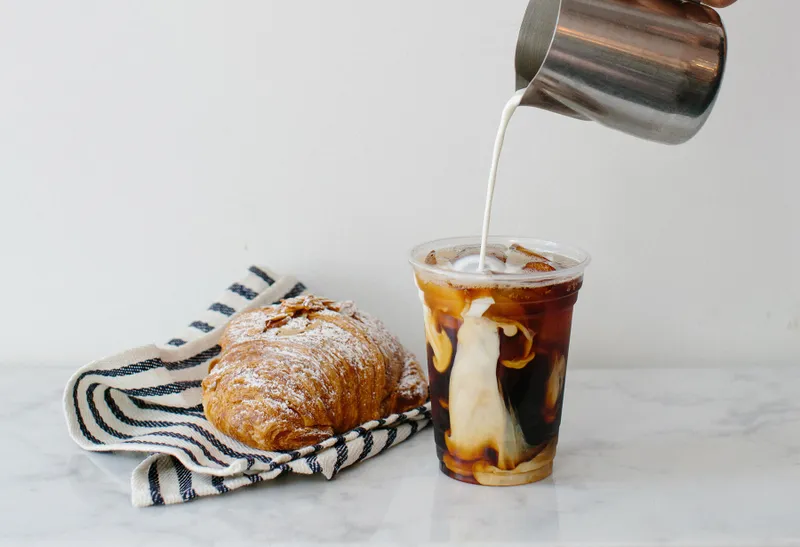 Iced coffee with milk being poured into it beside a croissant on a striped napkin on a marble surface.